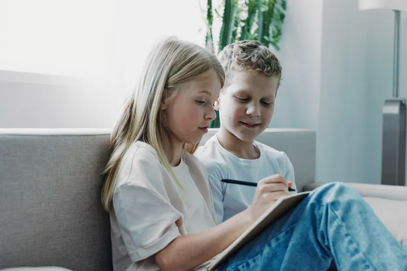 Two children sitting on a couch focused on drawing in a notebook together in a bright living room. Concept of creativity, learning, childhood, and friendship in a cozy home environment.
