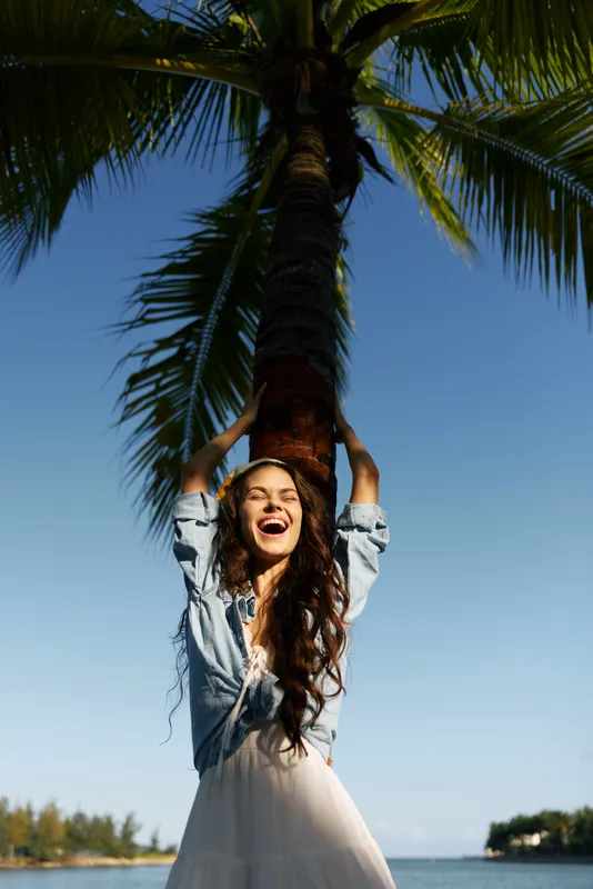 Smiling woman enjoying sunny day by palm tree near the sea with casual denim jacket and relaxed joyful mood.