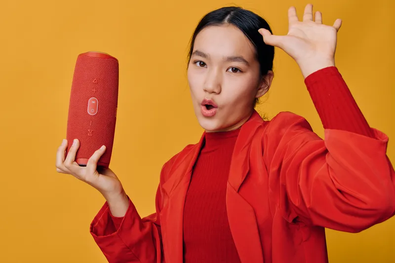 Young woman holding red wireless speaker and posing with open mouth on yellow background. Music, audio device and casual fashion with vibrant look for technology and entertainment.