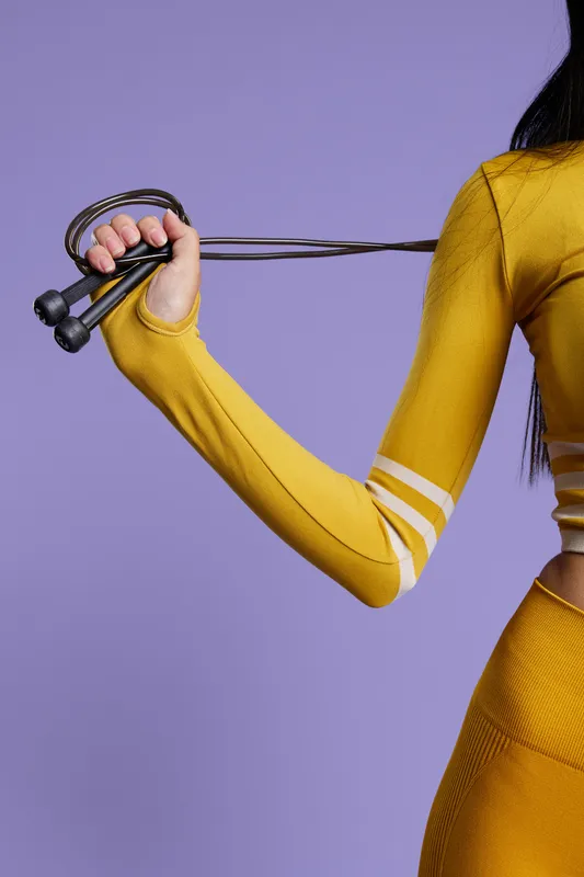 Woman sport fitness demonstrating jump rope exercise in bright yellow activewear, close-up of arm and torso with vibrant purple background, healthy lifestyle concept.