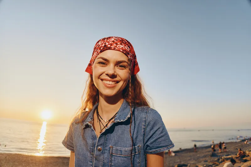 smiling woman with bandana on beach at sunset wearing denim shirt and natural makeup in warm golden light relaxing outdoors