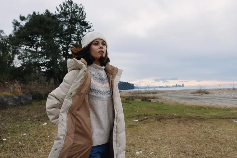 Winter outdoor portrait of a woman wearing a long coat and beanie, standing in a windy field by the water with a distant horizon and cloudy sky, conveying warmth, style and calmness in chilly weather.