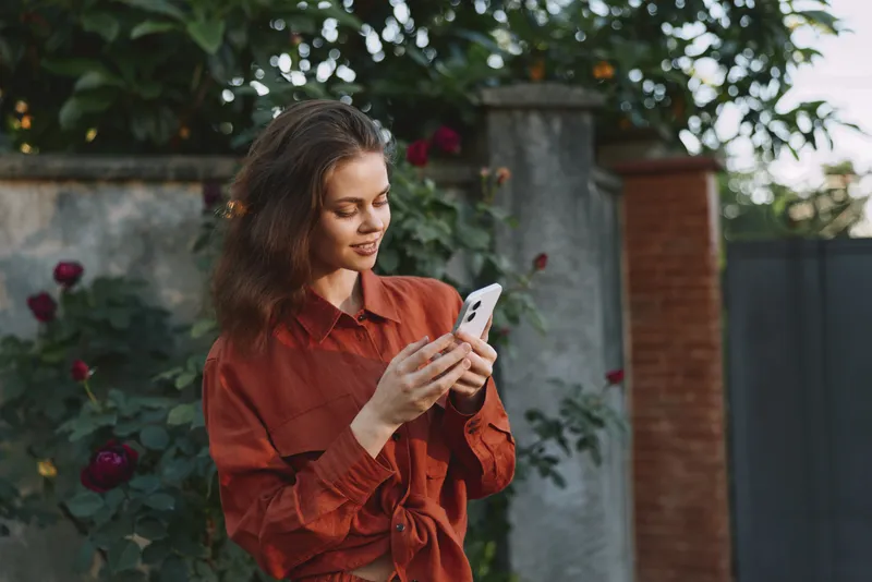 Young woman in an orange shirt stands in a sunlit garden, gazing at her phone with a soft smile, relaxed posture and warm natural light