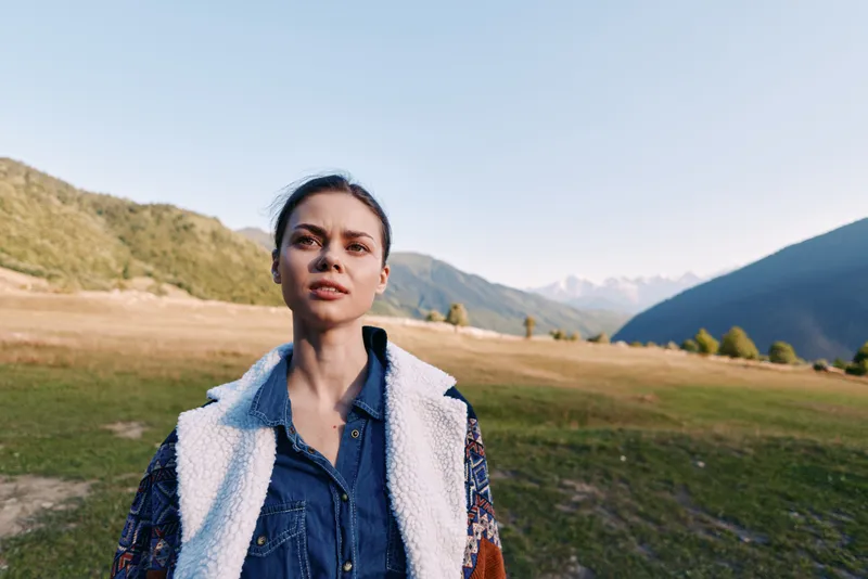 Woman portrait in mountains meadow nature outdoors, young female model wearing denim jacket with sherpa lining, standing in valley with serene sky and sunlight for travel lifestyle.