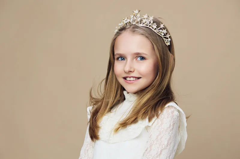 Charming portrait of a young girl wearing a tiara, smiling against a neutral beige background, showcasing a playful and whimsical atmosphere ideal for childhood memories