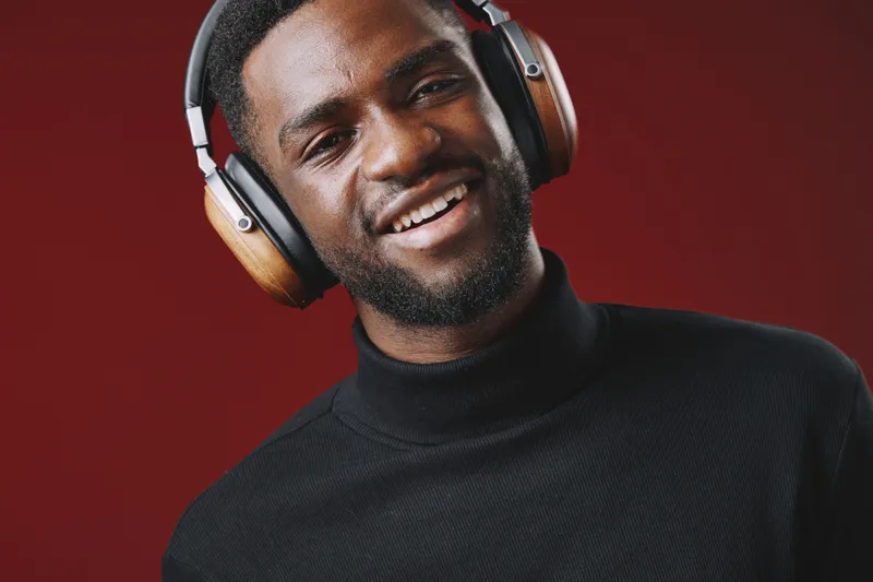 Young black man smiling while listening to music with headphones, wearing a black turtleneck against a warm red background, conveying joy and relaxation