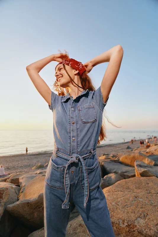 Lifestyle woman in modern boho-western denim street style enjoying sunset by the beach with a film color tone, smiling and relaxed in casual outfit.