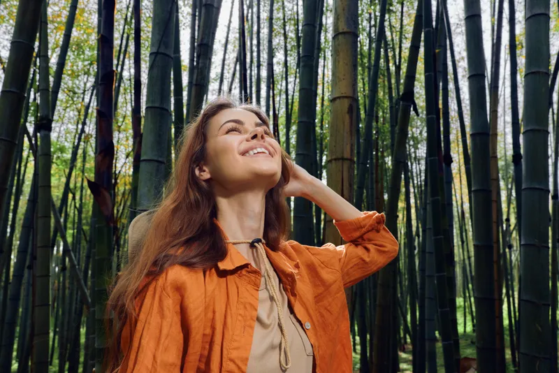 Woman in bamboo forest nature portrait smiling and looking up outdoors, relaxed pose amid tall bamboo trees, casual orange jacket and serene green environment with peaceful expression.