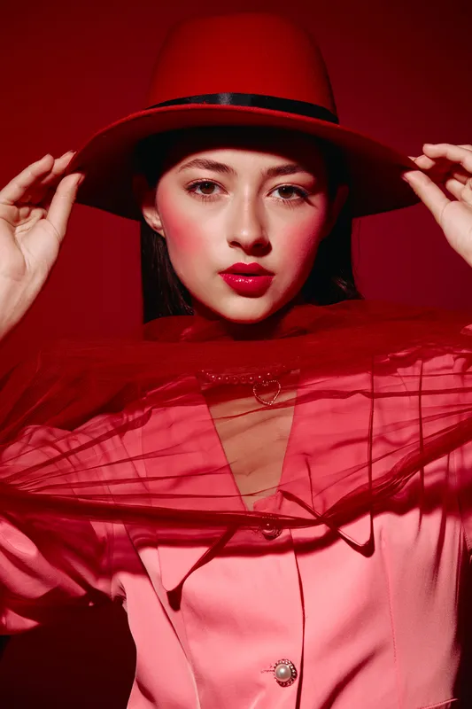 Stylish woman wearing a red hat and vibrant makeup poses against a red background. The sheer red fabric adds texture, highlighting beauty, fashion, and creative expression.