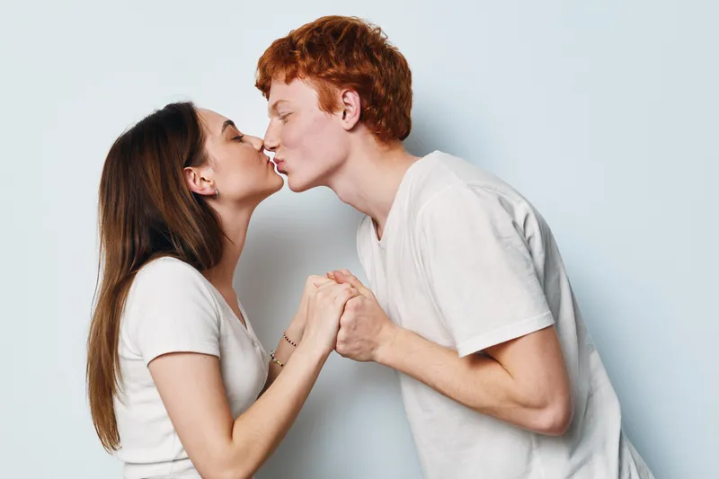 Young couple kissing with closed eyes holding hands in loving romantic moment. Man and woman in casual white t shirts expressing intimacy and affection. Studio portrait.