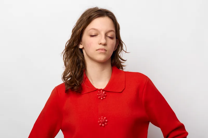 Young woman in red sweater with closed eyes and relaxed expression stands against white background. Concept of calm, meditation, mindfulness, and inner peace.