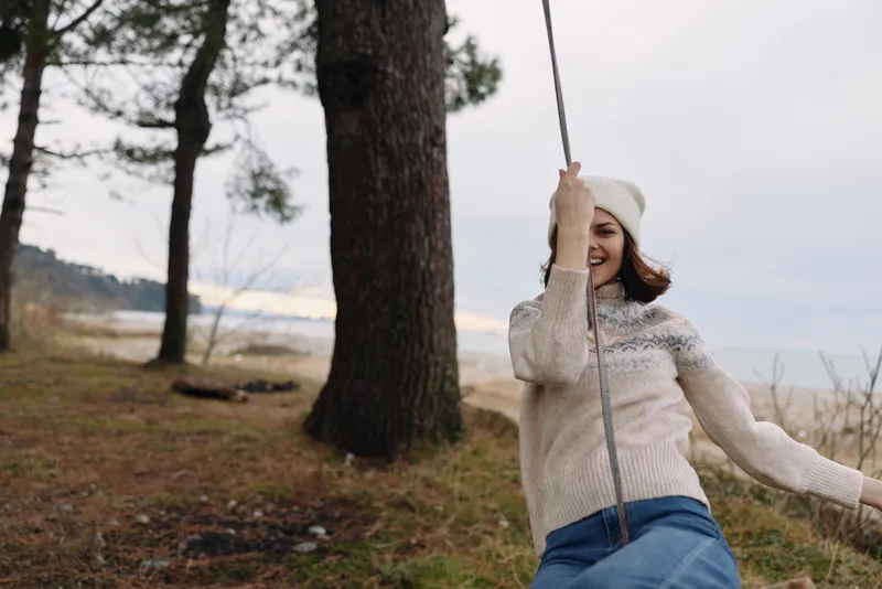 Woman on a swing outdoors wearing a knitted sweater and beanie, laughing among trees by a lake, creating a warm natural moment for a candid lifestyle shot