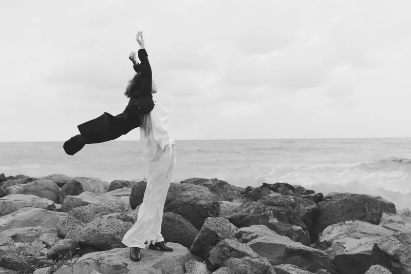 Couple on a rocky shore with arms raised in triumph, balancing on jagged stones as waves crash behind, a monochrome scene of motion, freedom and connection in an outdoor coastal landscape
