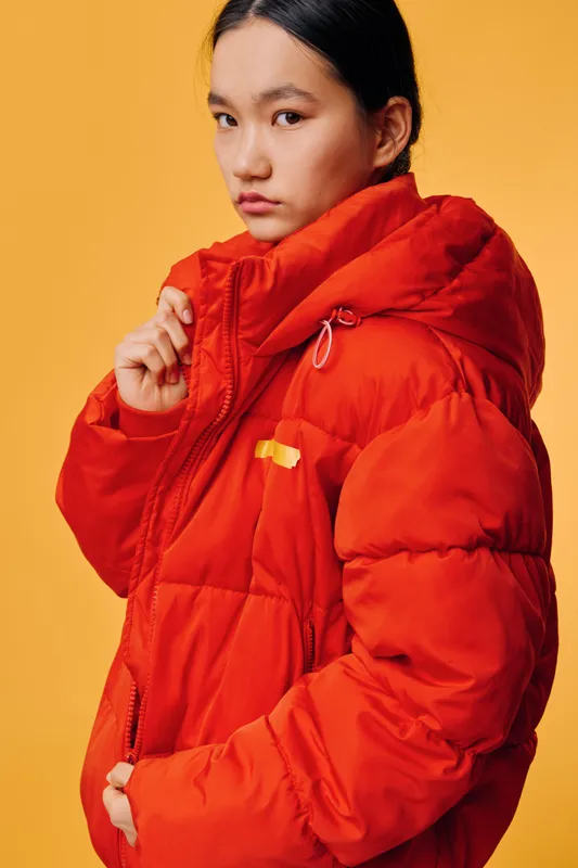 Young woman wearing red puffer jacket posing against yellow background with serious expression. Fashion portrait with warm clothing, winter style, and casual outfit for cold weather.