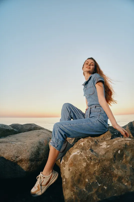 lifestyle woman wearing modern boho-western denim street style outfit sitting on rocks at sunset with film color effect and relaxed expression
