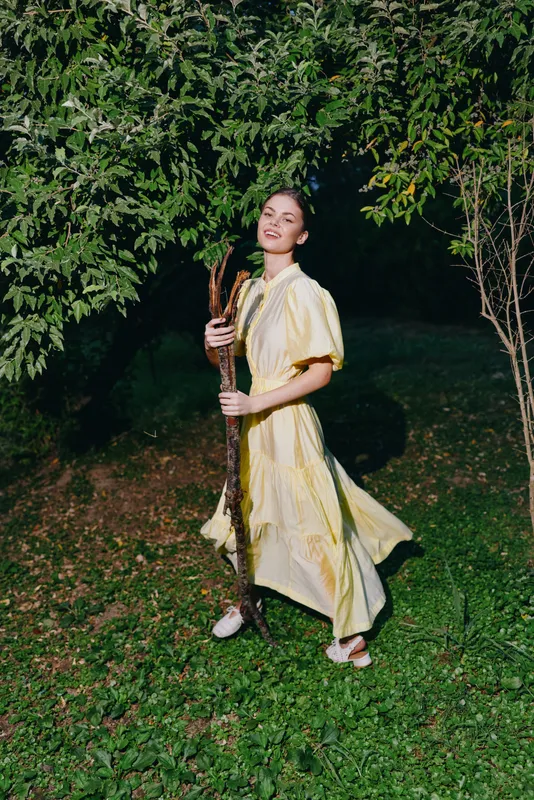 Woman in a pale yellow dress stands outdoors at night, smiling while holding a tree branch, surrounded by lush greenery and a soft mystic atmosphere