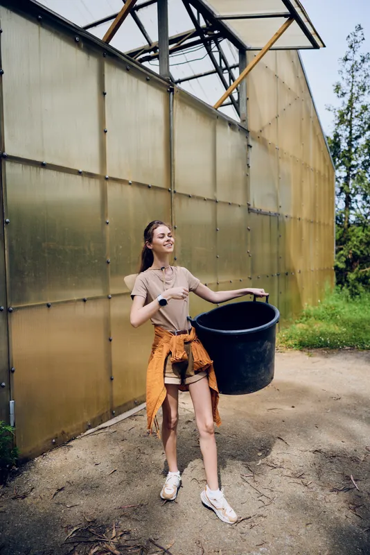 Woman bucket greenhouse gardening outdoor young female carrying a large tub by greenhouse, casual outfit with sneakers, smiling while moving soil and plants during summer activity.
