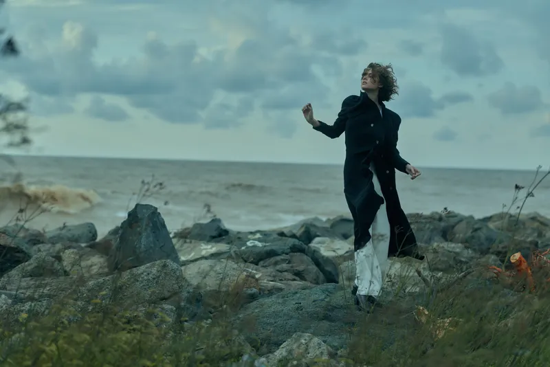 Coastline scene with a figure in a dark coat standing on rocks beside the sea, wind swept and dramatic, exploring rugged shoreline and outdoor fashion in a moody seascape