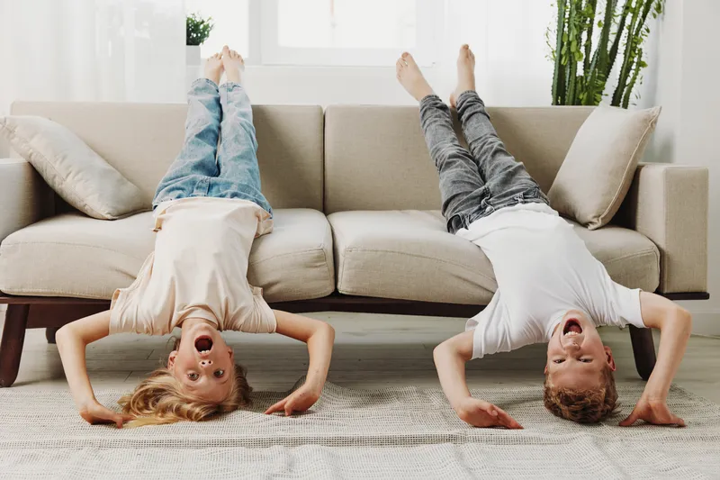 Two playful children doing headstands on the floor in a bright living room with a beige sofa and natural light.