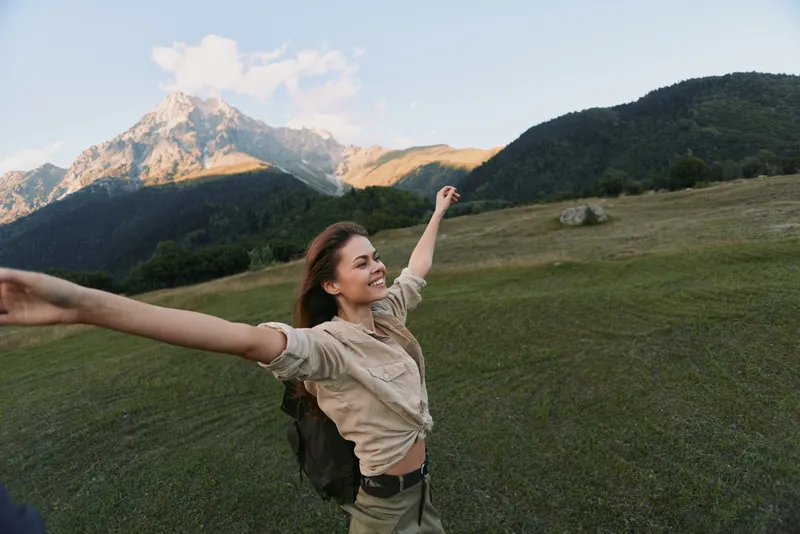 Young woman with long brown hair enjoys freedom with arms outstretched in a mountain meadow during a sunny day, expressing happiness and relaxation in nature.