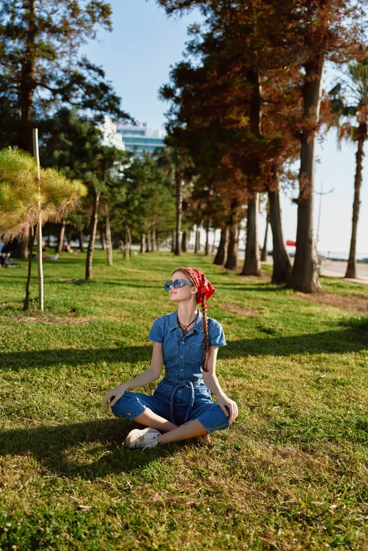 Lifestyle woman in modern boho-western denim street style sits cross-legged on grass wearing sunglasses and red headscarf in warm sunlight with film color tones.