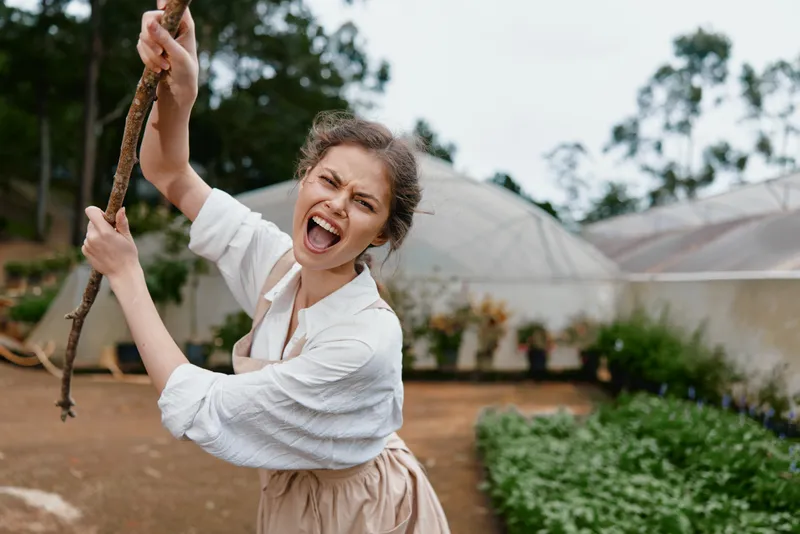 Young woman expressing joy while holding a stick in a garden, wearing casual farm style clothing amidst green plants and a natural background