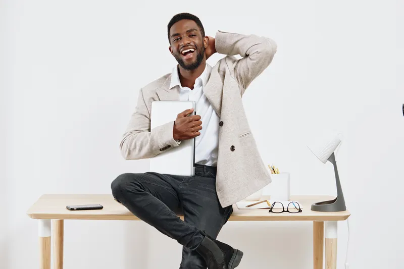 Smiling man in smart casual outfit sitting on desk in modern office interior holding tablet device, cheerful positive energetic professional lifestyle concept