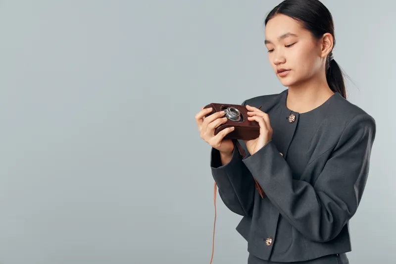Young woman wearing gray jacket holding vintage camera with focus at light gray background. Female photographer inspecting old camera for hobby or photo shoot with calm expression indoors.