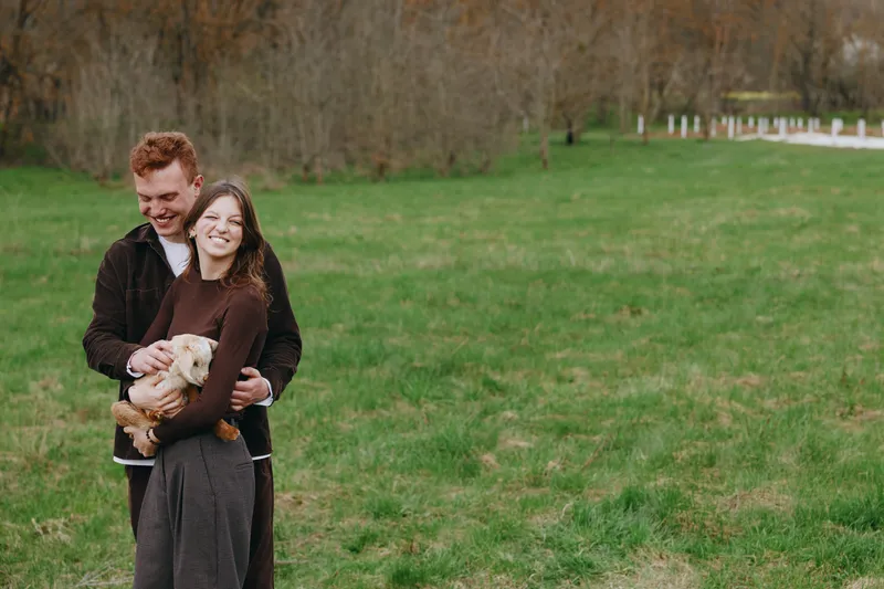 Couple outdoors in a park warmly embrace their small dog, smiling at the camera, enjoying a relaxed lifestyle moment in natural light, green grass, casual clothing, affectionate pose, candid family