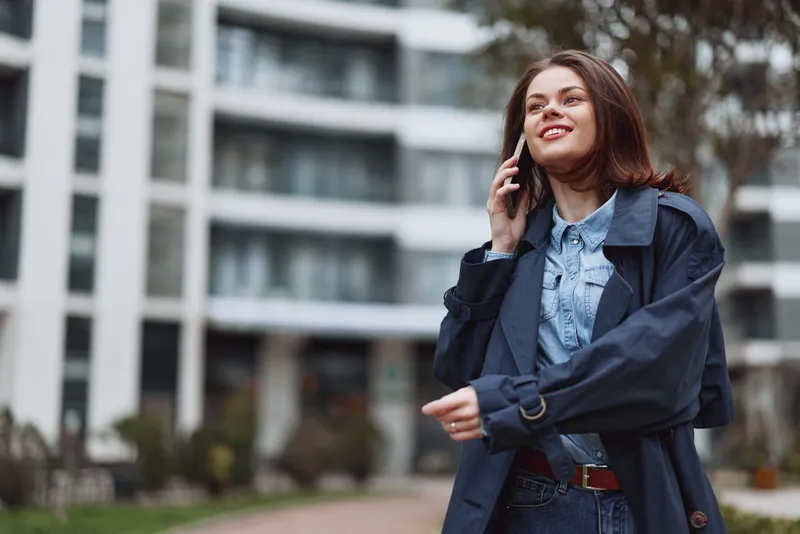 Young woman with long brown hair wearing a dark coat and denim shirt smiles while talking on a smartphone outdoors near modern buildings on a cloudy day.