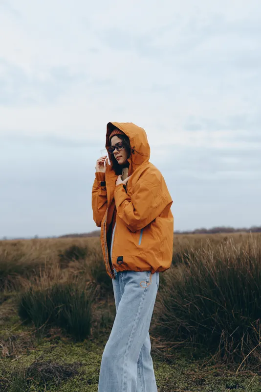 woman lifestyle outdoors hooded jacket field nature cloudy portrait standing, orange hooded coat and blue jeans, hood up in wind, grassy meadow background, soft daylight, peaceful mood