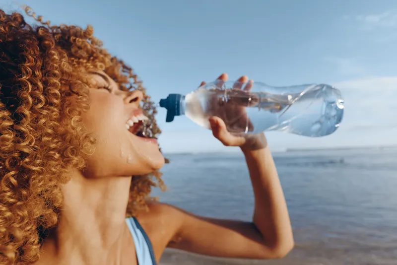 Young woman with curly hair drinks water from a plastic bottle on a sunny beach, enjoying a refreshing moment and feeling energized by seaside air, her athletic vibe suggesting a workout break