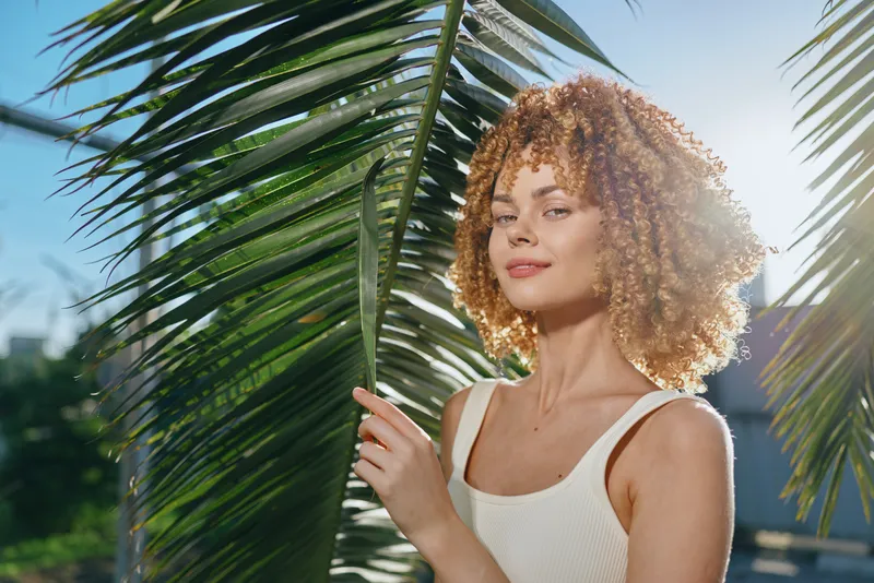 Smiling woman with curly hair stands near tropical palm leaves in warm sunlight wearing white tank top, radiating natural beauty and relaxed summer vibe outdoors.