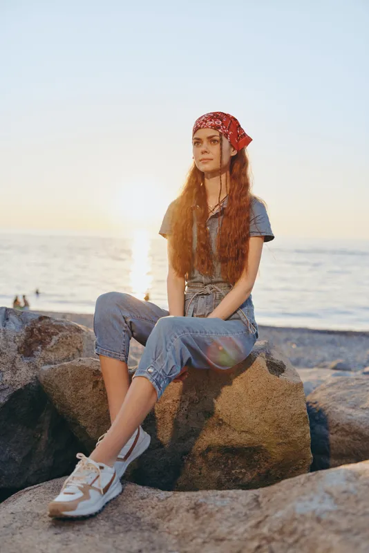 lifestyle woman in modern boho-western denim street style sitting on rocks by the sea with warm film color tones and natural sunset light