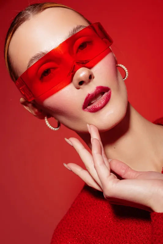Close-up portrait of a stylish woman wearing red transparent sunglasses and red lipstick. She poses with hand near face against a vibrant red background. Fashion and beauty concept.