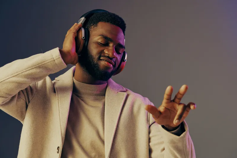 A young African man enjoying music, wearing stylish headphones and a beige coat, set against a colorful gradient background, exuding happiness and relaxation