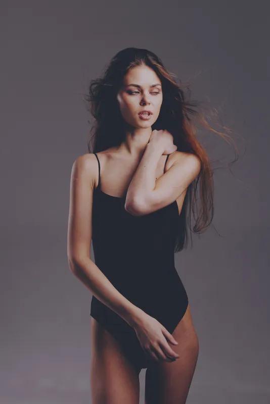 slender woman in black bodysuit posing with thoughtful expression and tousled hair on gray background