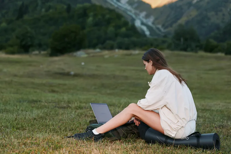 A lone individual sits on a mat in a grassy field, typing on a laptop. She wears light clothing and appears focused, enjoying a peaceful outdoor work moment.