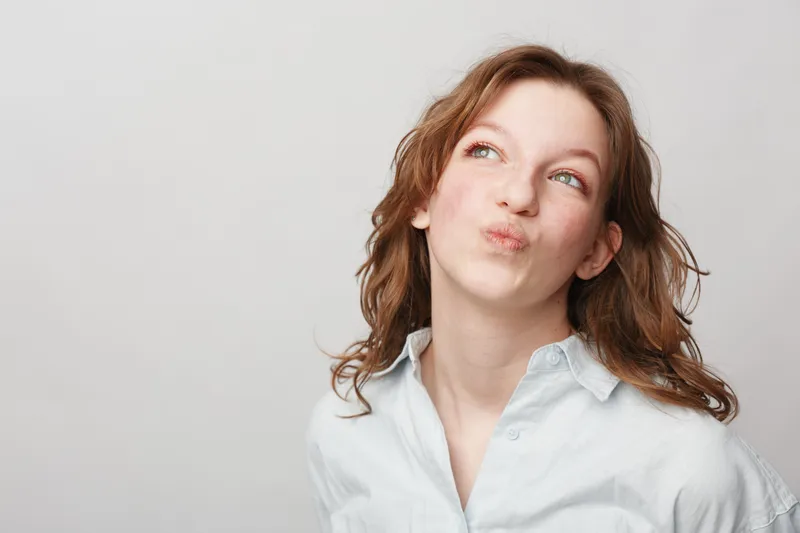 Young woman with curly hair wearing white shirt looks up thoughtfully with pursed lips. Studio portrait on light background. Concept for thinking, curiosity, and contemplation.