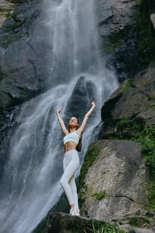 young woman practicing yoga near waterfall in white activewear, outdoor fitness, meditation, nature, healthy lifestyle, serenity, athletic pose