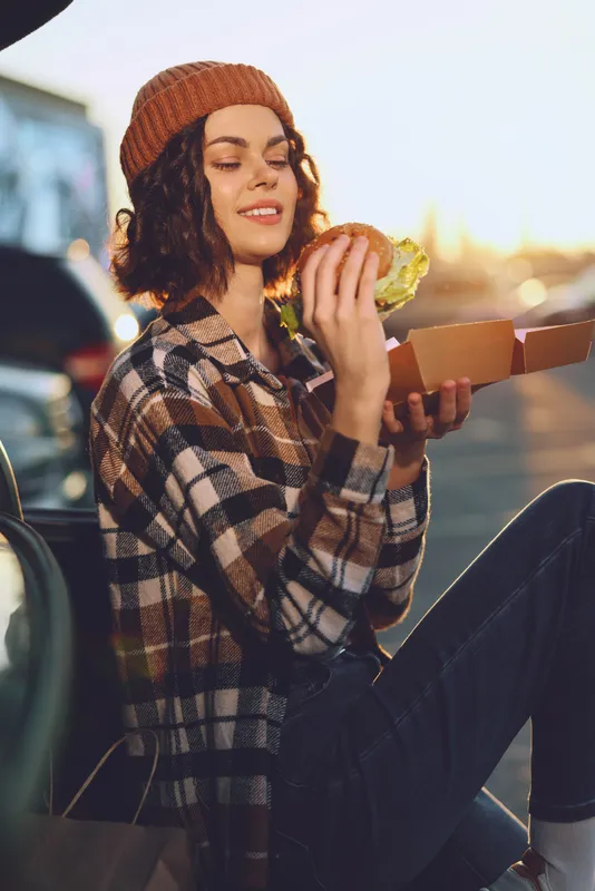 woman burger smile beanie car sunset plaid Young woman in a plaid jacket smiles while eating a burger from a box in a car at sunset, golden hour glow, authenticity, candid lifestyle, mindful living