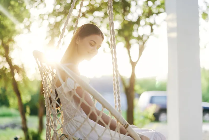 Beautiful young woman in white dress sitting in a hanging chair, enjoying a peaceful moment with sunlight filtering through trees, embodying serenity and tranquility