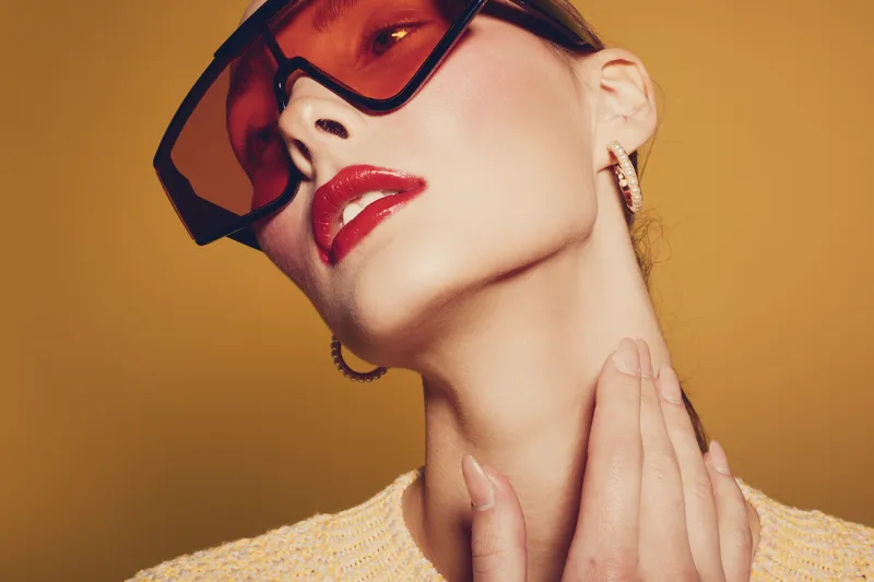 Close-up of stylish woman with red lipstick and oversized sunglasses touching neck. Fashion portrait with warm yellow background and soft lighting. Concept of beauty and elegance.