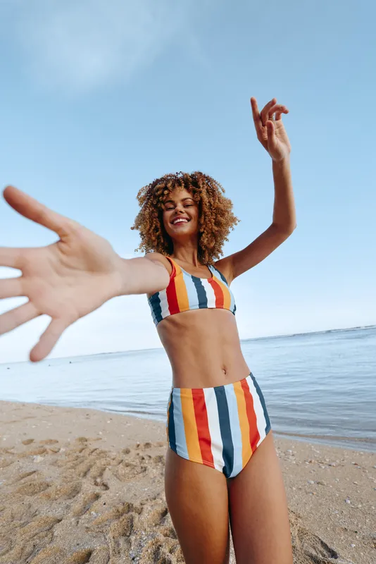 A young woman in a colorful striped bikini on a sunny beach reaches toward the camera with a wide smile, the ocean and blue sky forming a bright backdrop.