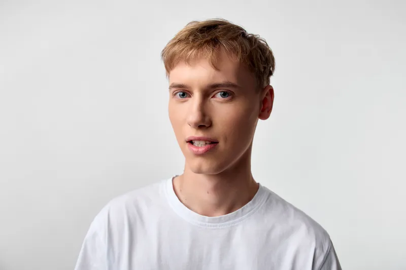 Young man in a white t shirt stands against a plain backdrop with natural lighting, showcasing casual style and a neutral expression for commercial, ecommerce and fashion projects