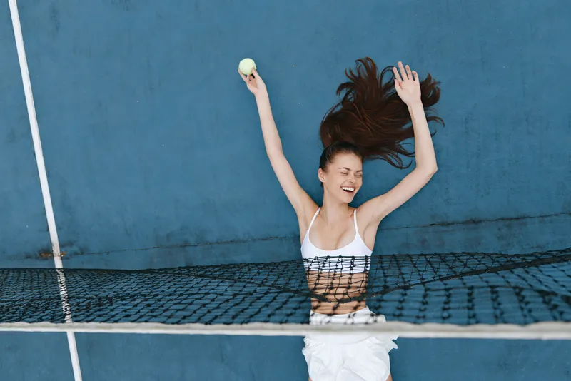 Joyful young woman celebrating after winning a tennis match, wearing a sporty white outfit, with a bright blue background, perfect for sports and fitness themes