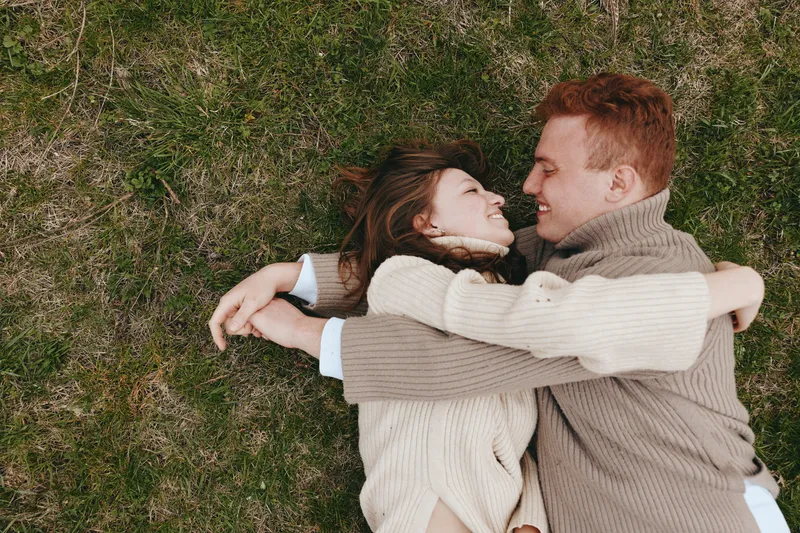 A couple lying on grass, embracing and smiling in an outdoor setting. A candid lifestyle moment showing warmth, affection, and connection in a relaxed, natural environment, perfect for love and