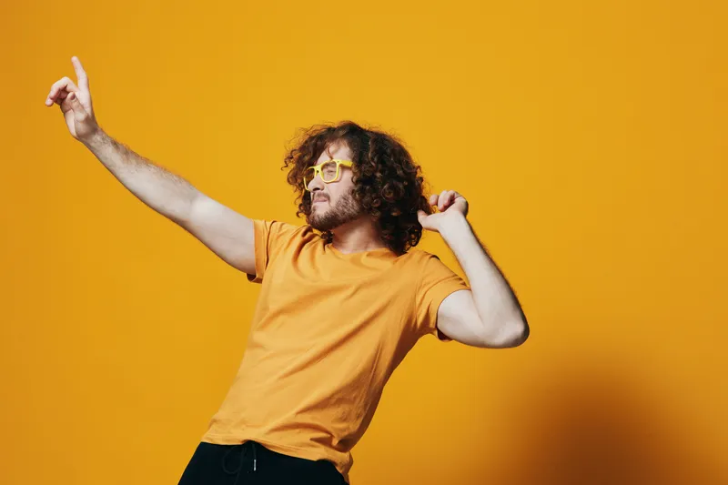Happy young man with curly hair wearing yellow glasses and a T shirt, dancing joyfully against a vibrant yellow background, radiating positive energy