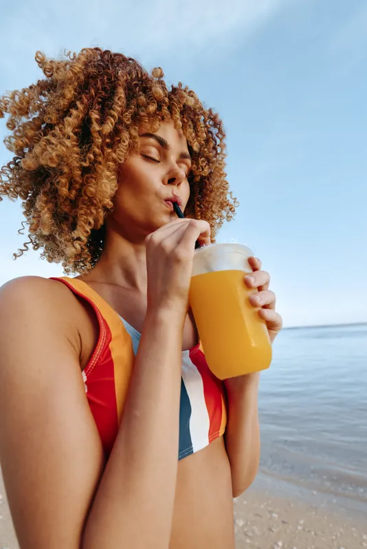 Young woman with curly hair enjoys a fresh orange juice by the beach on a sunny day, wearing a colorful striped swimsuit and feeling relaxed and refreshed.