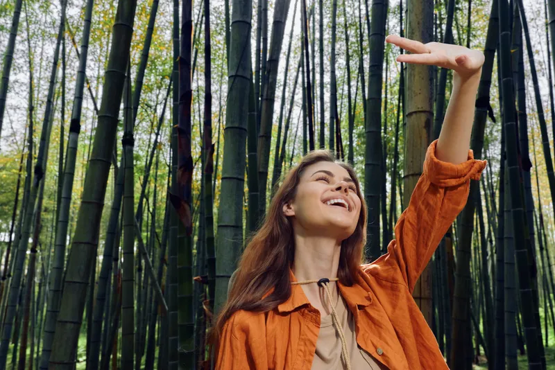Woman smiling portrait in bamboo forest, nature scene with tall trees and outdoors atmosphere. Female traveler in orange jacket raises hand shading eyes while enjoying greenery.
